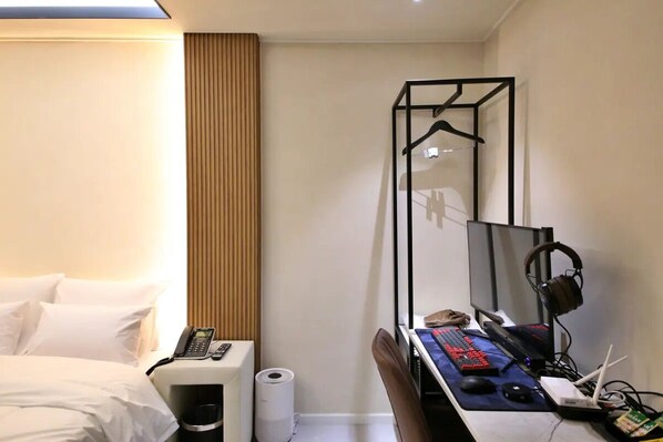 Habitación de hotel con cama, escritorio con computadora, silla, perchero y ventana con persianas.