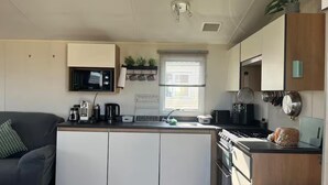 A compact kitchen with a microwave, kettle, toaster, and stove. There is a window with blinds, a plant, and a small shelf with kitchen utensils.