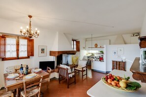 Un comedor con una mesa de madera, sillas y una araña. Se observa una cocina al fondo, con armarios y una encimera.