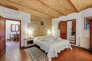 Un dormitorio con pared de piedra, techo de madera, cama con ropa blanca, alfombra estampada y un área de comedor visible a través de una puerta abierta.