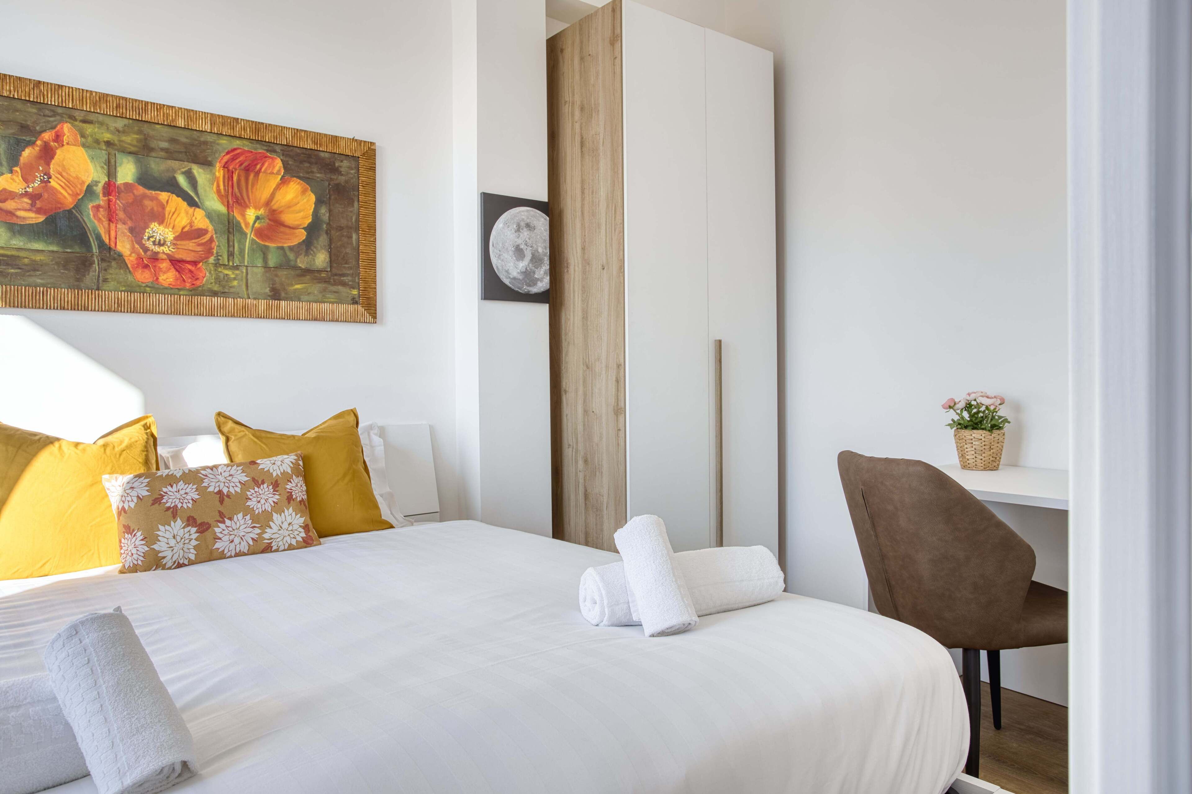 A neatly made bed with white linens, a brown floral pillow, and a white towel. A wooden panel divides the room, and a small shelf with a plant is visible.