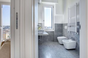 A modern bathroom with a toilet, bidet, and sink, featuring grey tiles and a window.