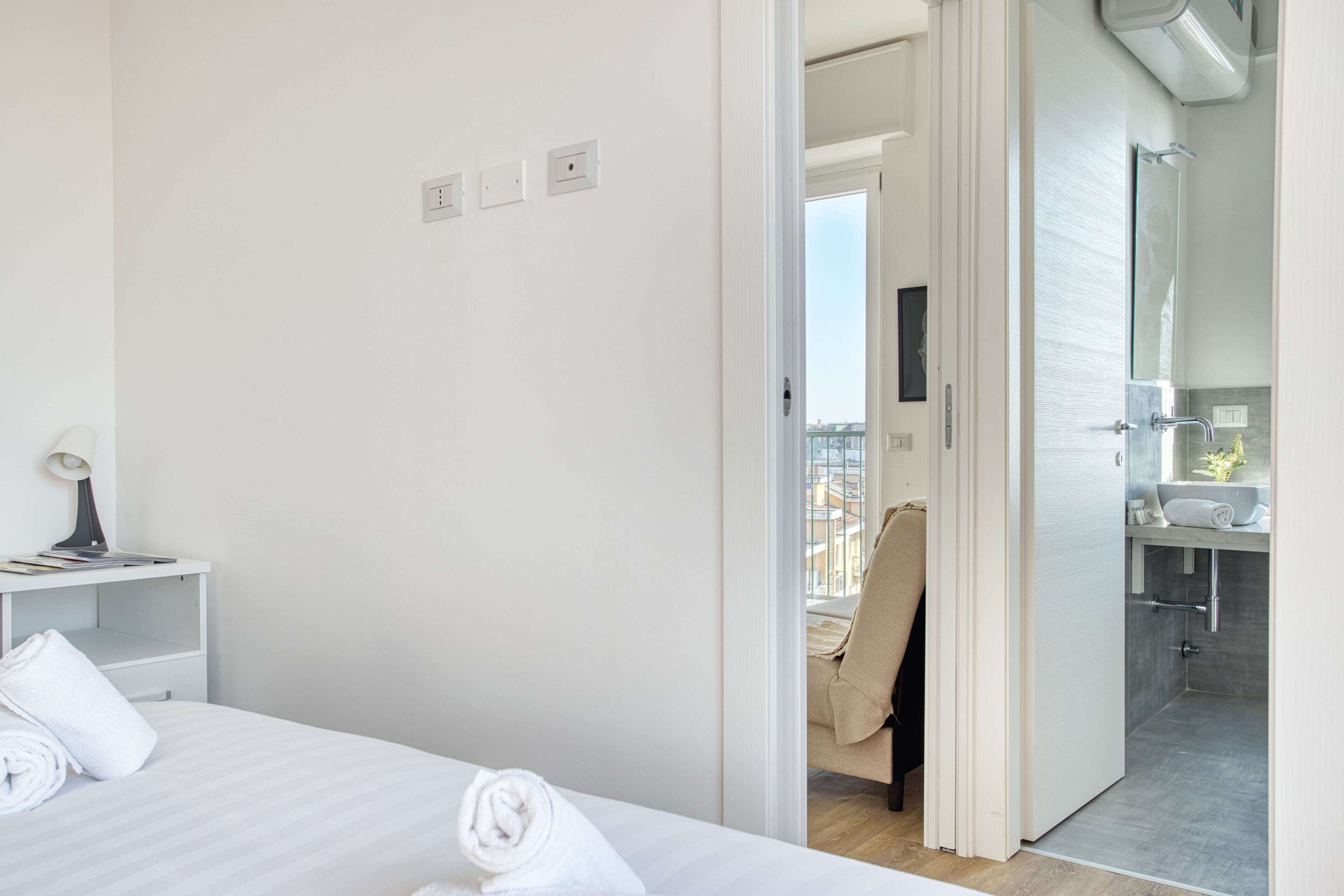 Moderni hotellihuone, jossa on siististi pedattu sänky, pieni työpöytä lampun kanssa sekä näkymä parvekkeelle ja kylpyhuoneeseen avonaisen oven kautta.