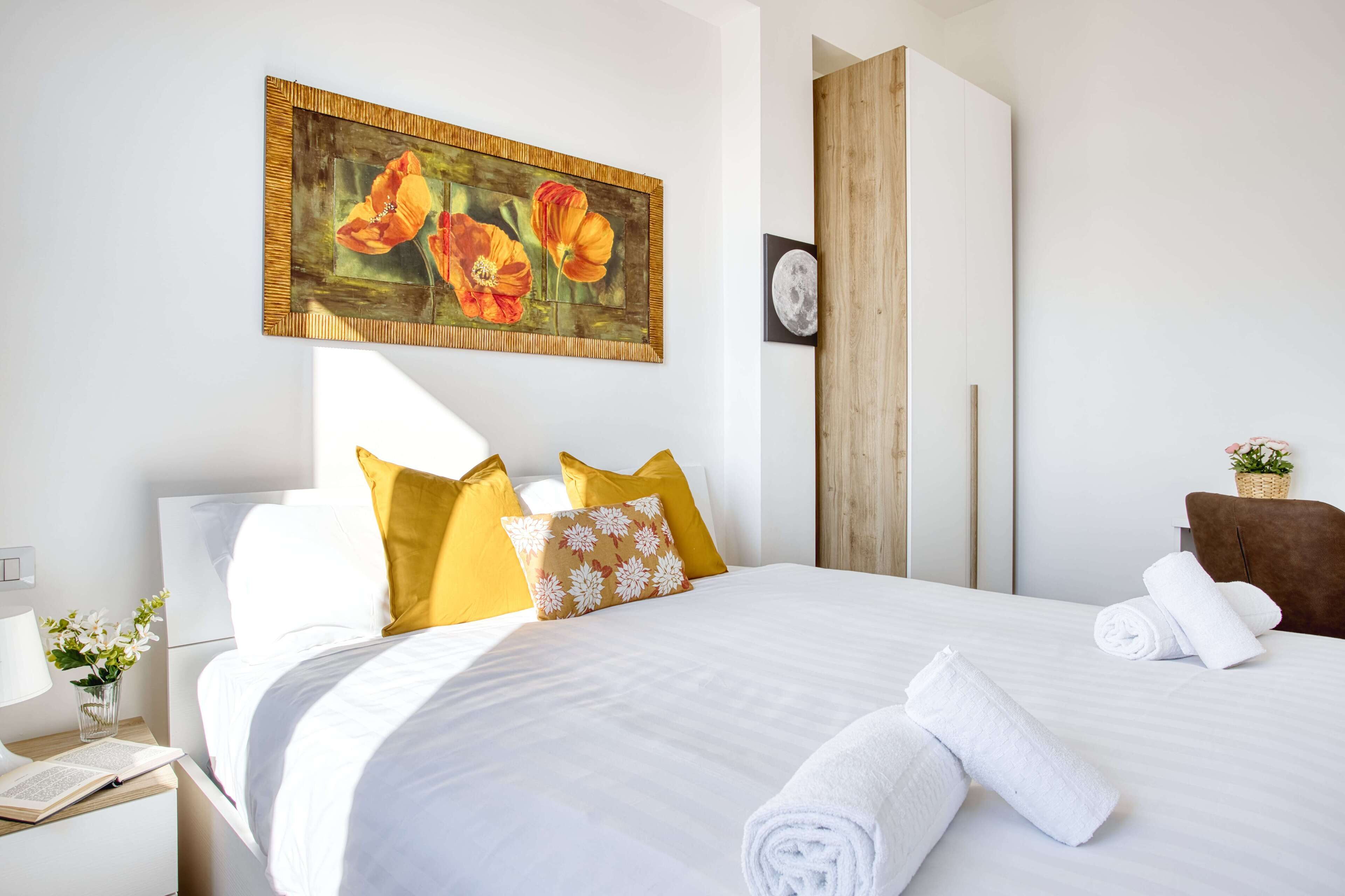 Un lit bien fait, avec du linge de lit blanc et des coussins jaunes, un tableau de fleurs accroché au mur et une armoire en bois.