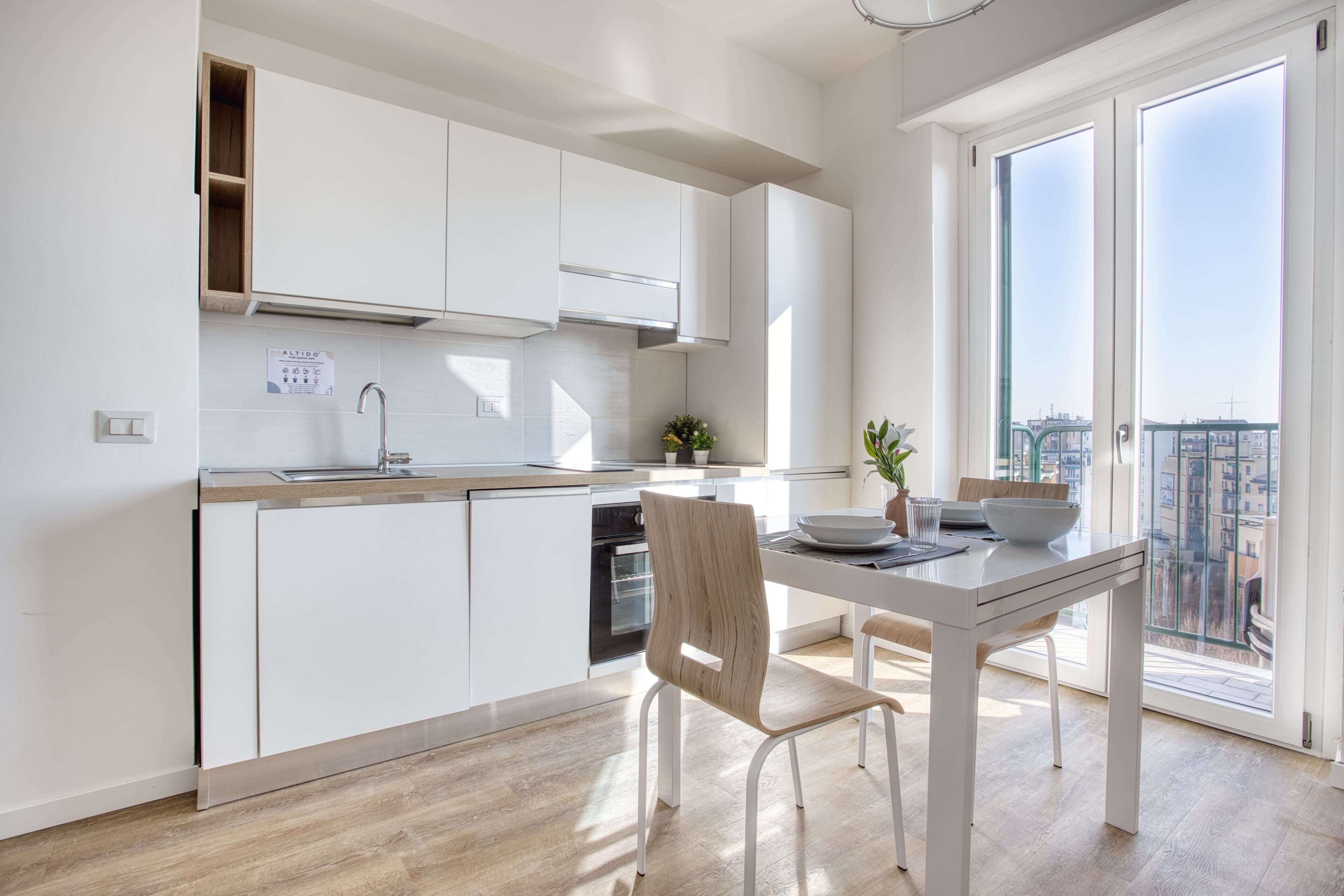 A modern kitchen with white cabinetry, a dining table with chairs, and a view of the city through large windows.