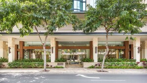 Una entrada de hotel moderna con fachada de vidrio, un área de acceso techada y un paseo arbolado.