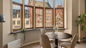 Una habitación con una mesa redonda y cuatro sillas, amplios ventanales con vistas a edificios rojos, una planta en maceta y una lámpara de pie.