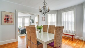 Uma sala de jantar com mesa branca, cadeiras de vime, lustre e janelas amplas com cortinas leves.