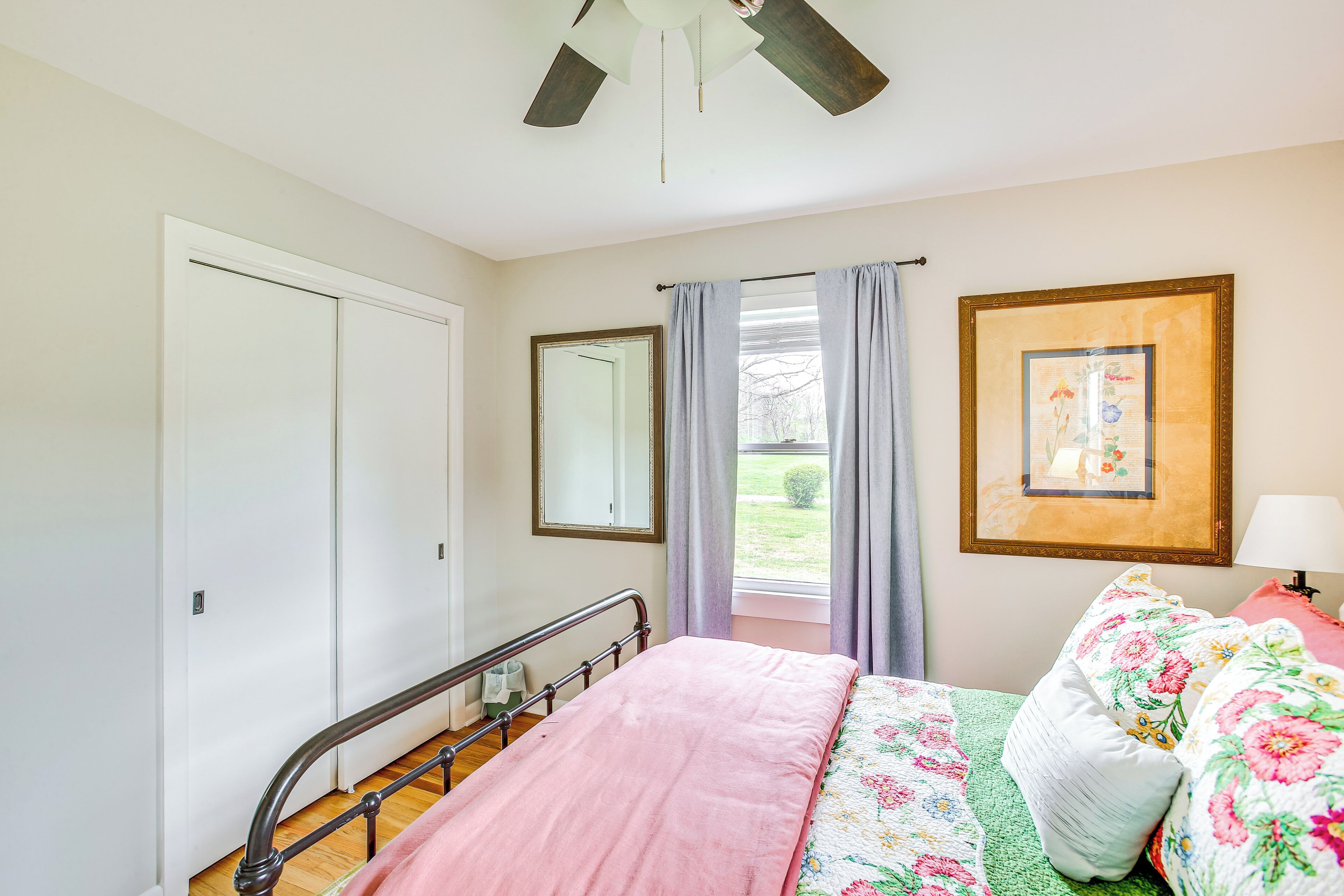 A bedroom with a bed, a ceiling fan, a window with curtains, a framed picture, and a bedside table with a lamp.