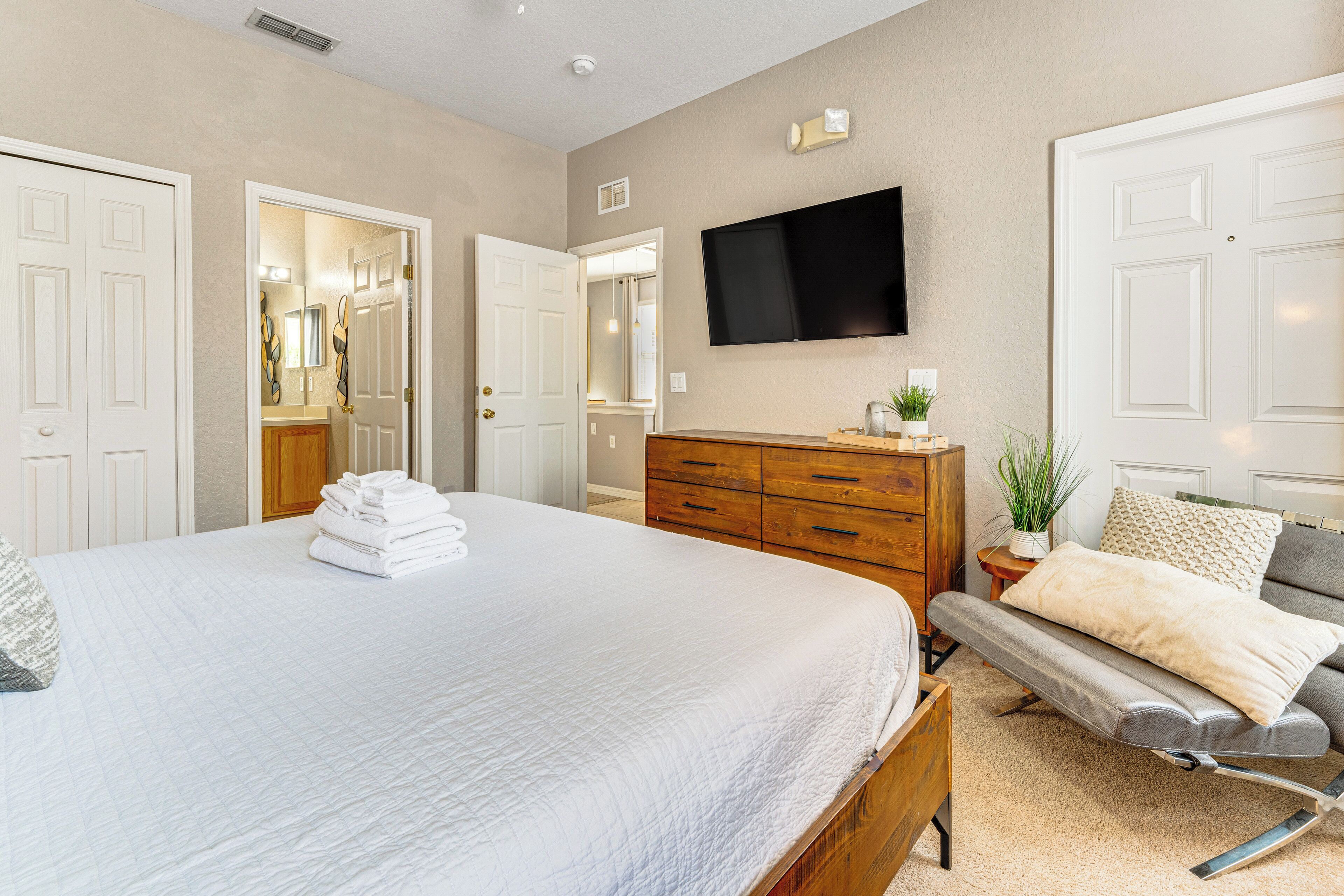 A bedroom with a large bed, a wooden dresser, a TV mounted on the wall, and a door leading to another room.