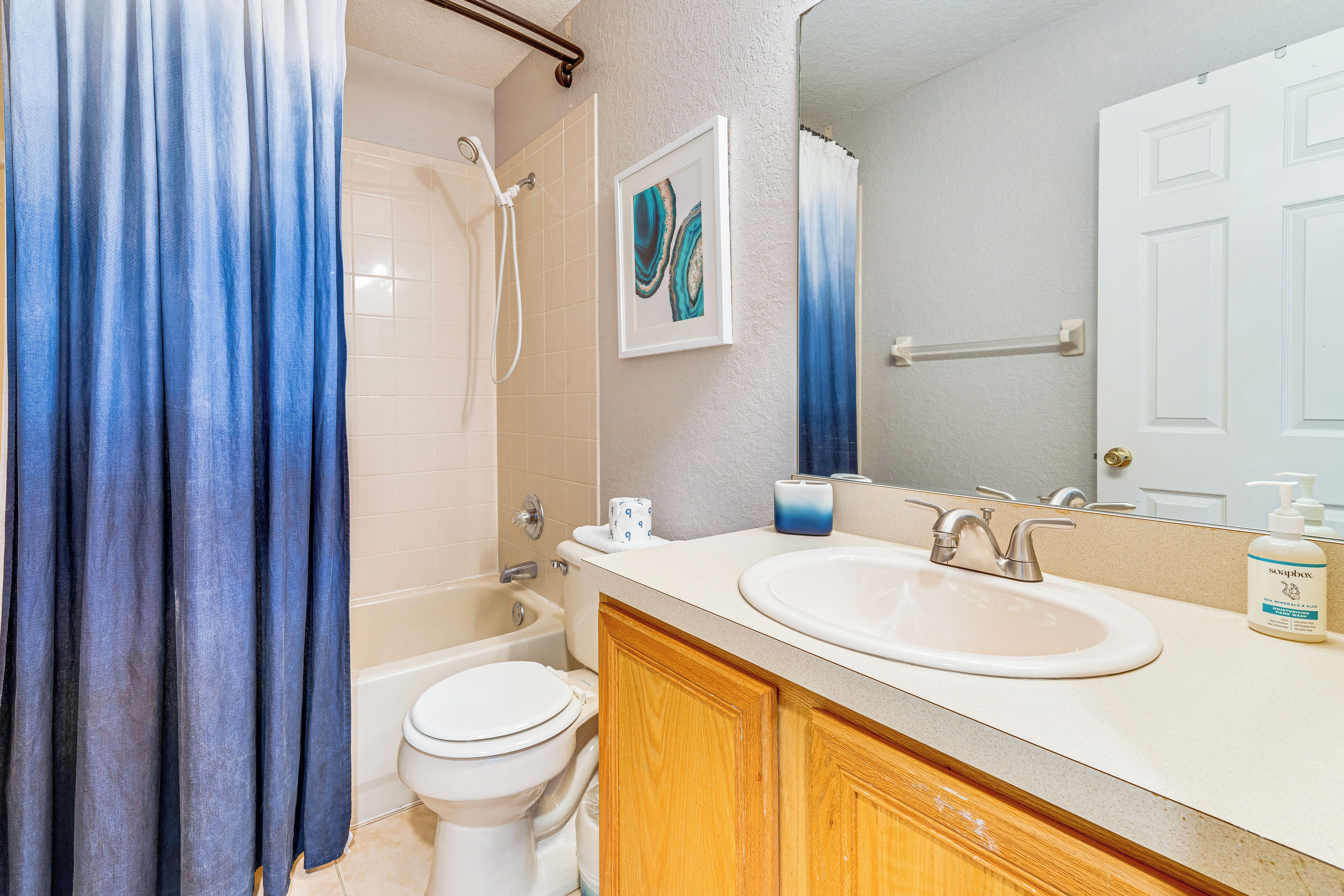 A bathroom with a bathtub, toilet, and sink. There is a shower curtain, a framed artwork, and a bottle of soap on the sink.