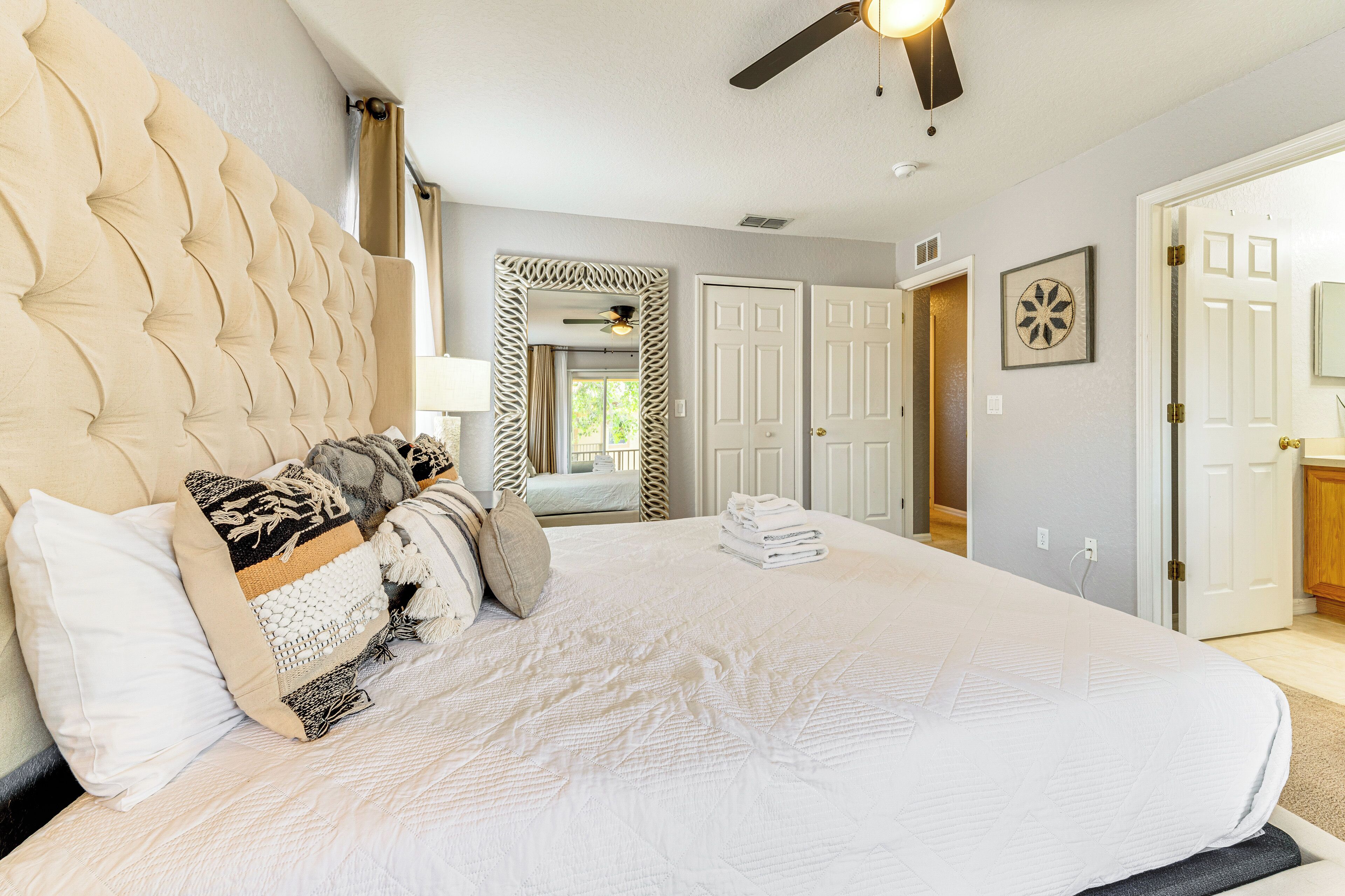 A large bed with a tufted headboard, a bedside table, and a mirror. There are two doors and a window in the background.