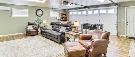A spacious living room with a large leather sofa, a brown leather armchair, a wooden coffee table, and a rug. There is a clock on the wall and a garage door in the background.
