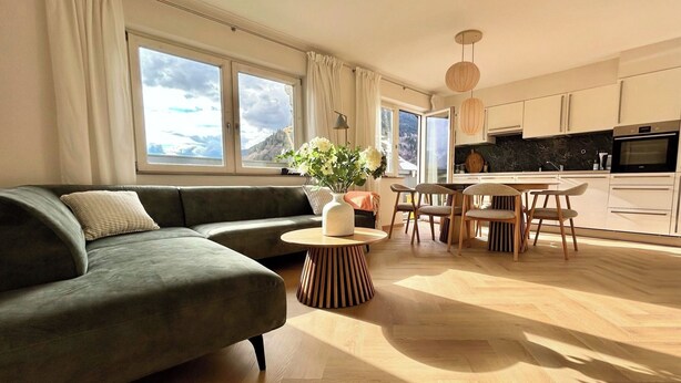 Ein modernes Wohnzimmer mit einer Couch, einem runden Holztisch und einem Essbereich mit Stühlen. Große Fenster mit Vorhängen, eine Küche mit weißen Schränken und eine hängende Leuchte.