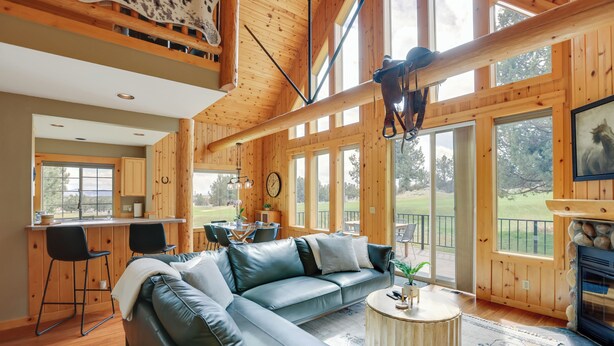 A spacious living room with a large leather sectional sofa, a round coffee table, and a fireplace. There is a kitchen area with wooden cabinets and a dining table. Large windows provide ample natural light and views of the outdoors.