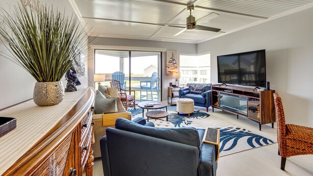 A spacious living room with a large TV, comfortable seating, a ceiling fan, and a view to the outside. - Beach Access: Ocean-view Condo w/ Screened Balcony (Harbor Island)