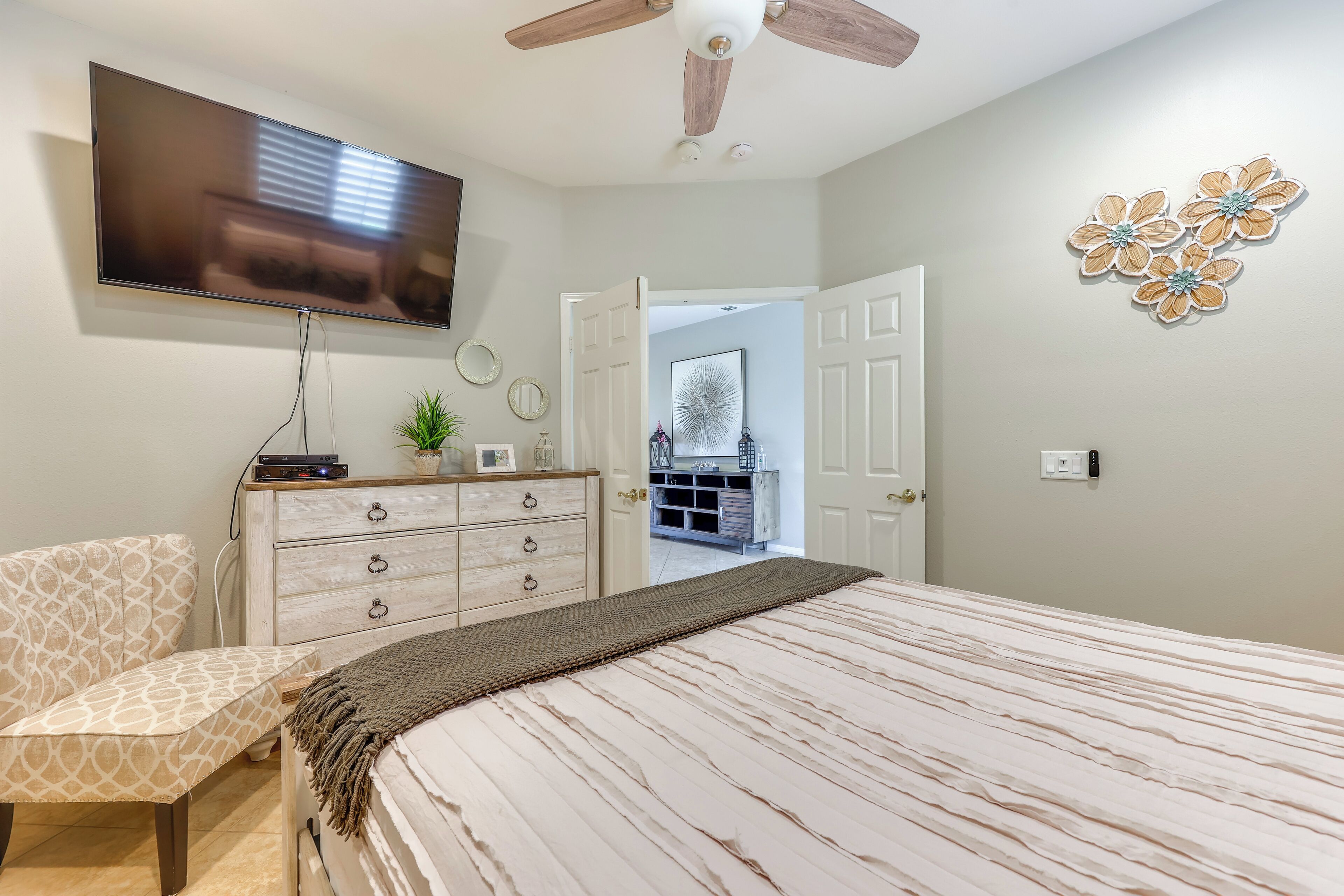 Une chambre à coucher comprenant un lit, un téléviseur, un ventilateur de plafond, une commode et une porte donnant sur une autre pièce.