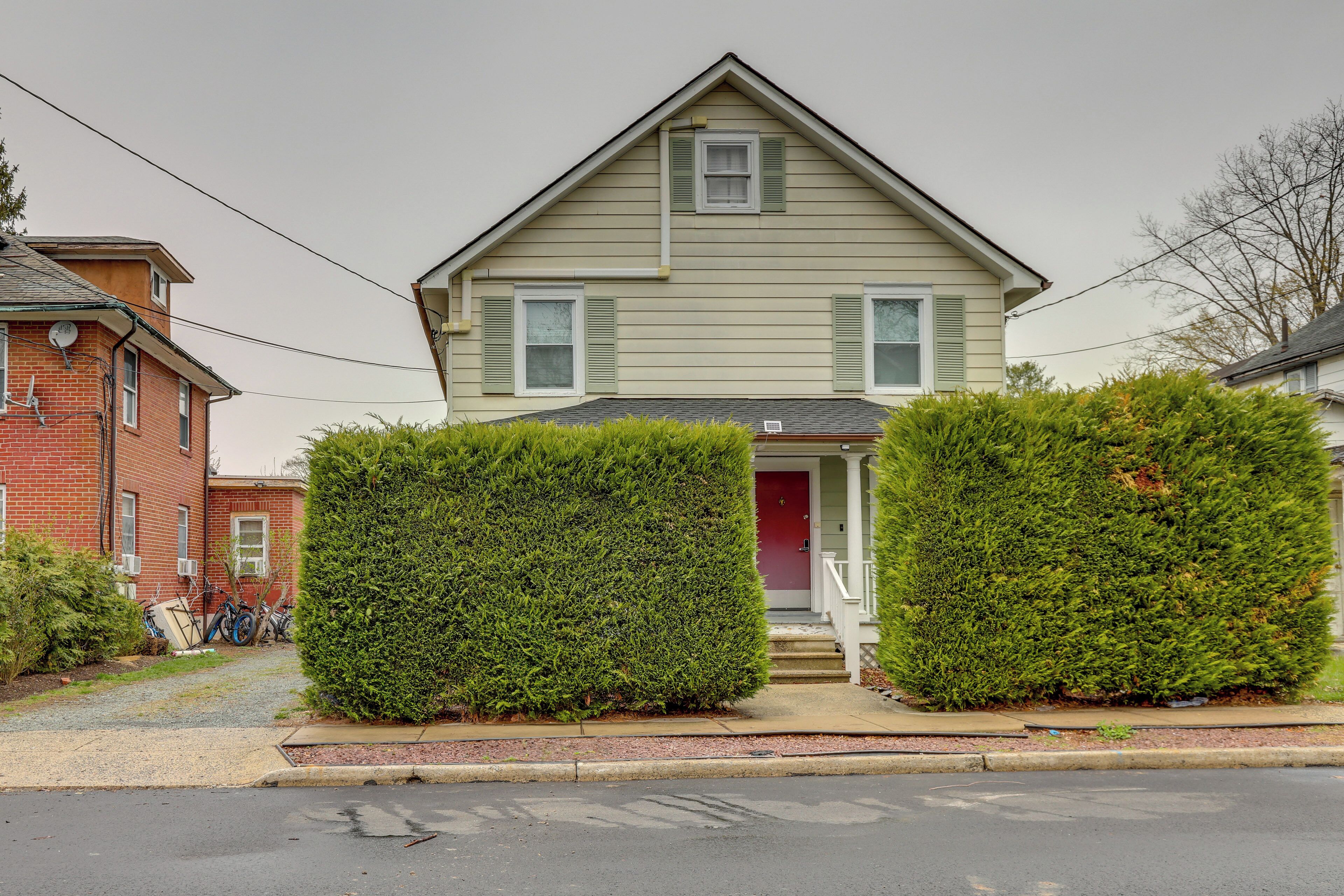 A two-story house with a red door and a small front porch. There are two large green hedges in front of the house.