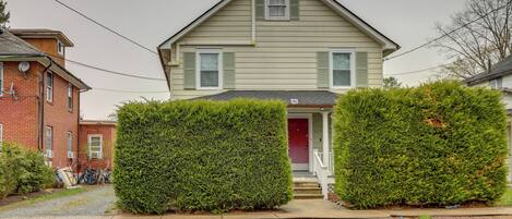 A two-story house with a red door and a small front porch. There are two large green hedges in front of the house.