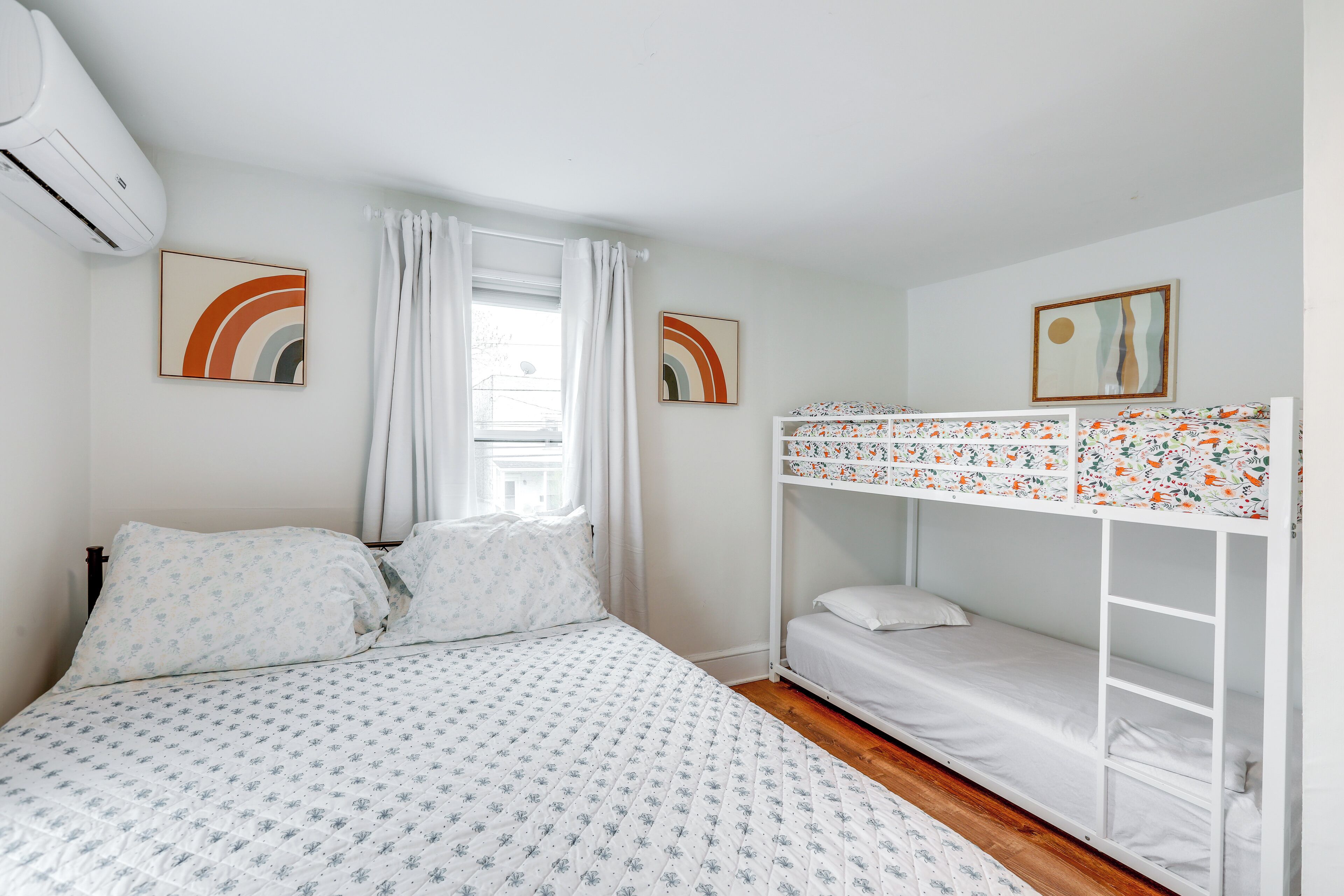 Een slaapkamer met een stapelbed, een bed met bloemenbeddengoed, een raam met gordijnen en twee kunstwerken aan de muur.
