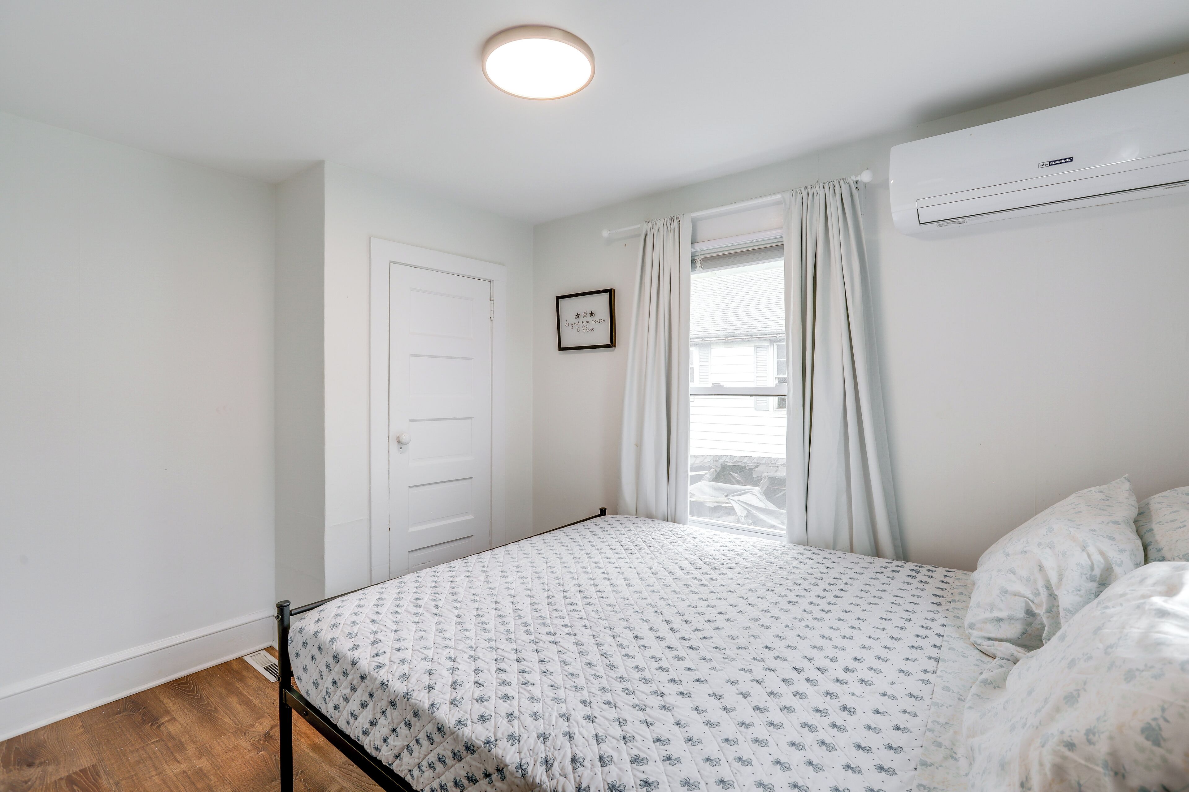 A bedroom with a bed, a window with curtains, an air conditioning unit, and a door.
