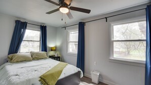 Une chambre à coucher avec un lit, un ventilateur de plafond, deux fenêtres avec des rideaux bleus, une table de chevet avec une lampe et une poubelle.