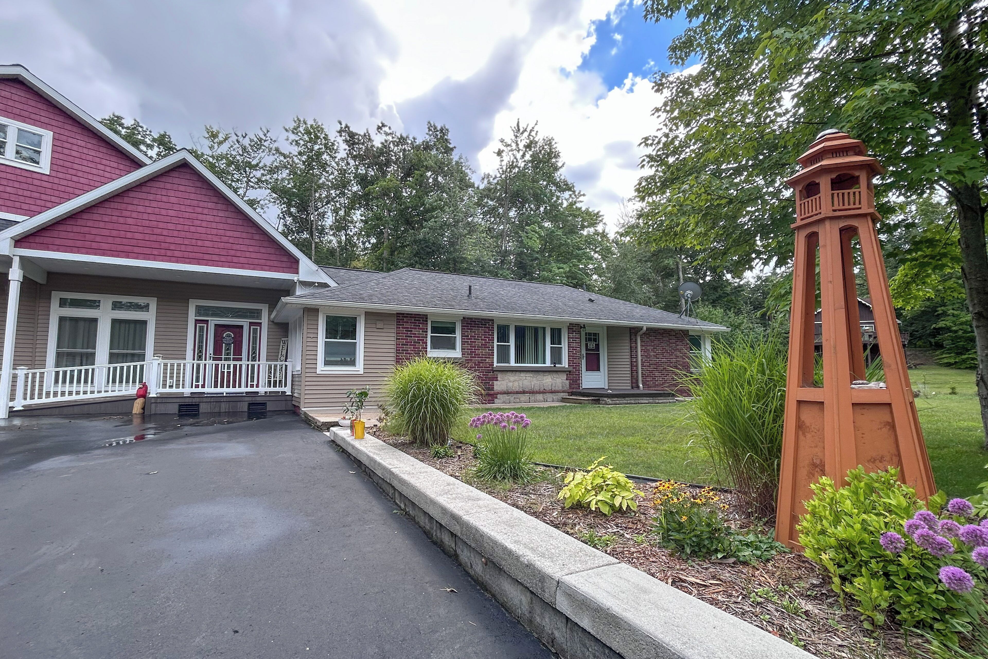 A two-story house with a red roof and beige siding, a front porch with a white railing, and a small garden with various plants and flowers.