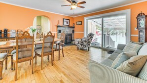 A living room with a wooden floor, a ceiling fan, a fireplace, and a large window. - Walk to Lake Huron Beaches: Home w/ Fire Pit! (Rogers City)