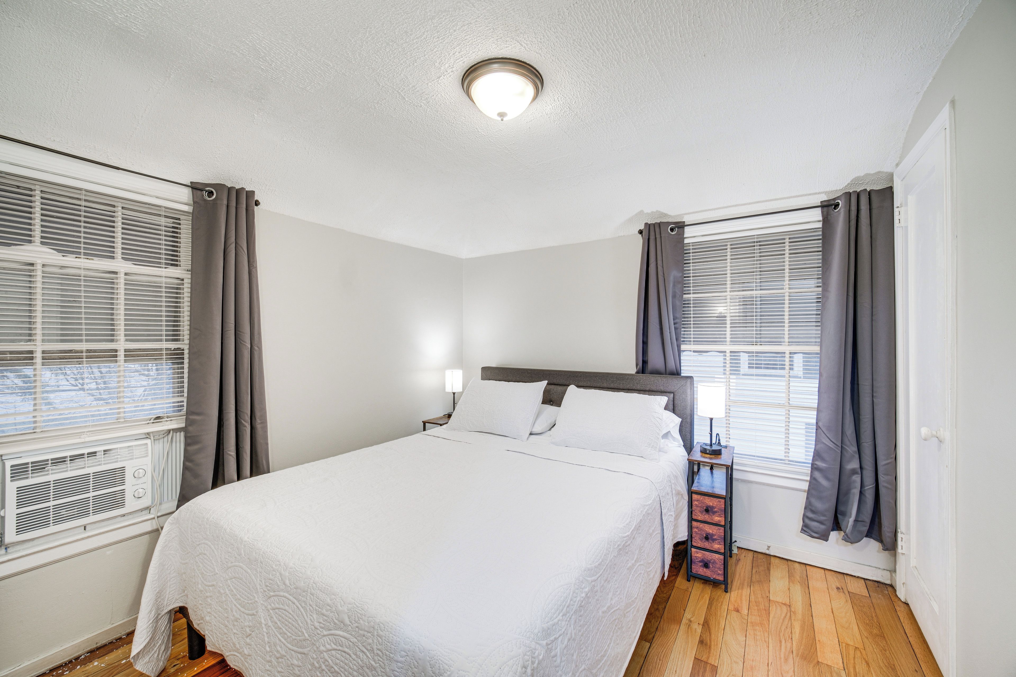 Un dormitorio ordenado con una cama grande, una mesita de noche, una lámpara y una ventana con cortinas.