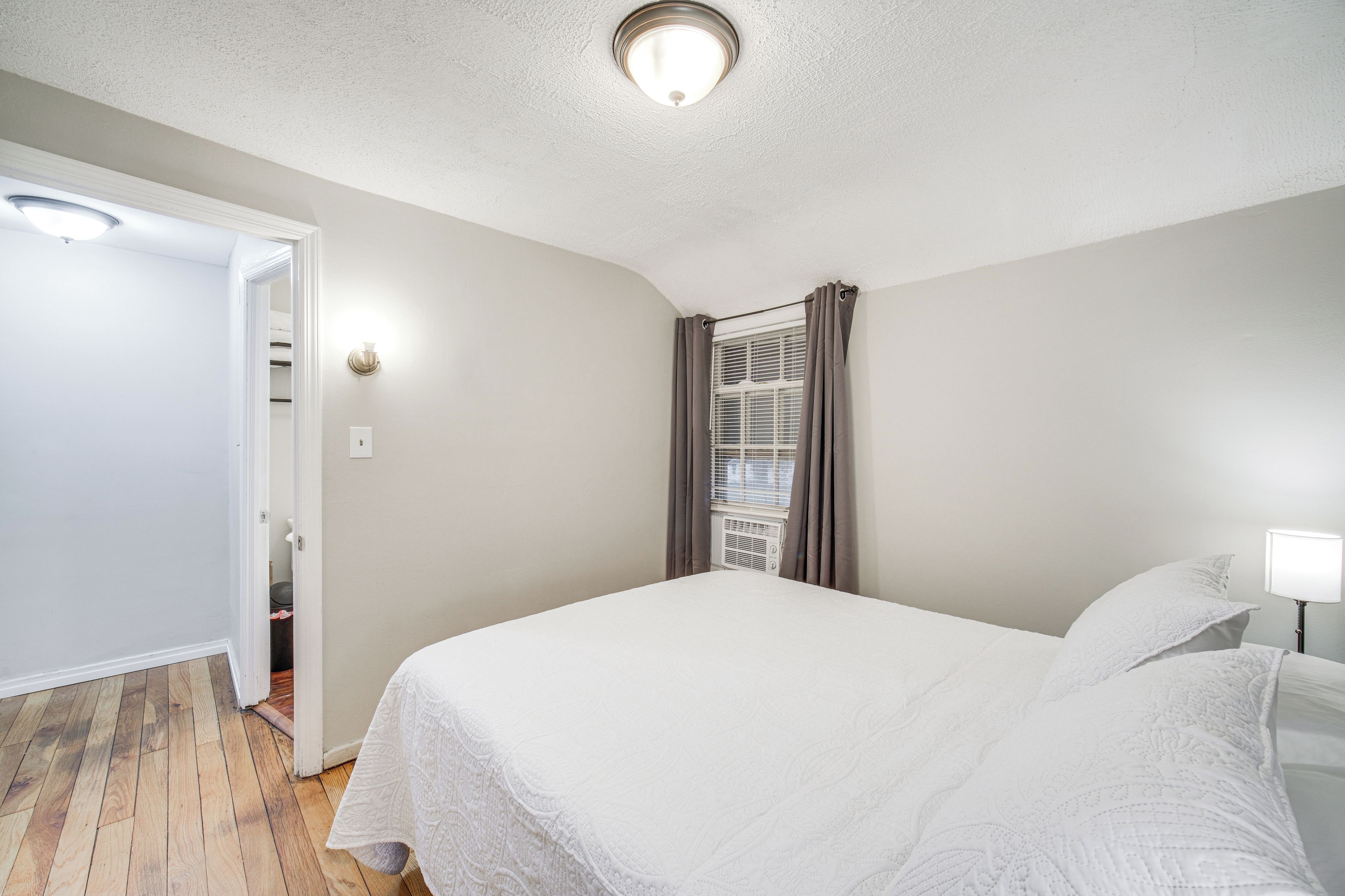 A bedroom with a bed, a window with curtains, a ceiling light, and a door leading to another room.