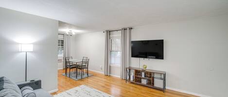 A modern living room with a grey sofa, a flat-screen TV, a dining area with chairs, and a wooden floor.