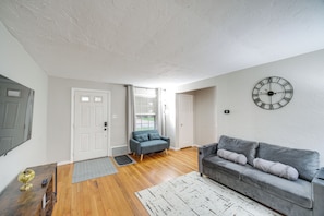 A living room with a grey sofa, a clock on the wall, and a wooden floor. - 2 Mi to Lake Erie: Home w/ Yard in Euclid! (Euclid)