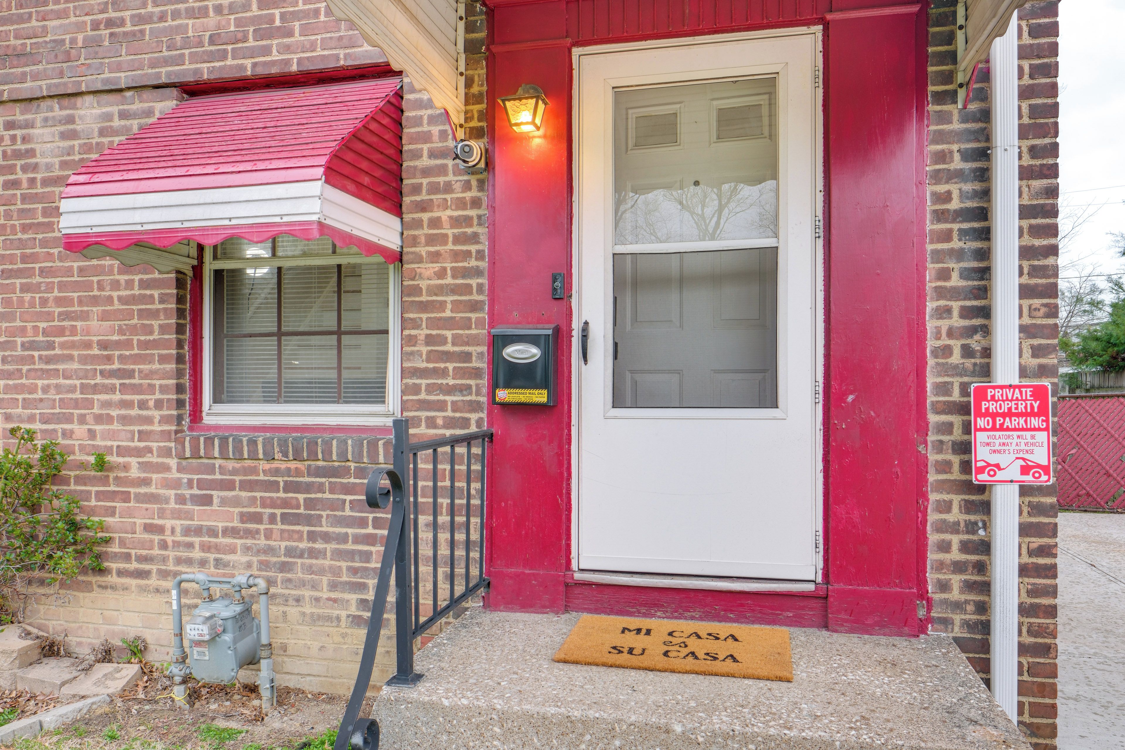 A red brick building with a white door, a red awning, and a doormat that reads "MI CASA SU CASA".