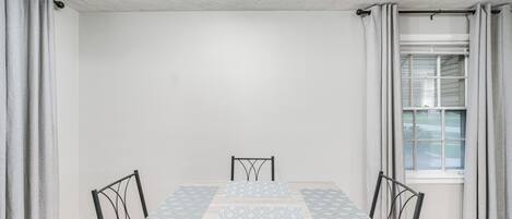 A dining room with a table and chairs, a window with curtains, and a white ceiling.