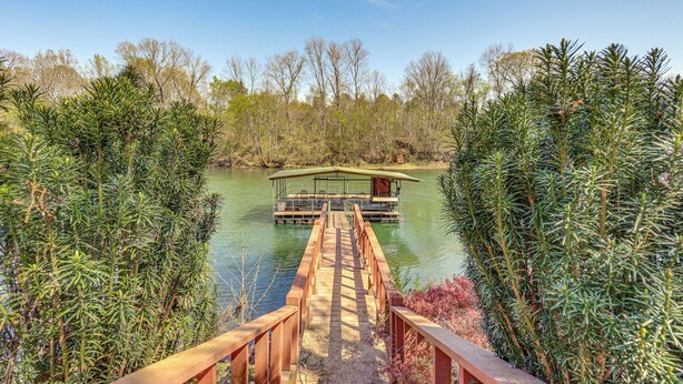 A wooden bridge leading to a floating structure with a covered deck and railings, surrounded by dense greenery and a calm body of water. - Private Dock: 'the River House' in Heber Springs! (Heber Springs)