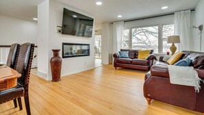 Een ruime woonkamer met houten vloeren, een bruine leren bank, een eettafel met stoelen, en een groot raam dat voor natuurlijk licht zorgt.