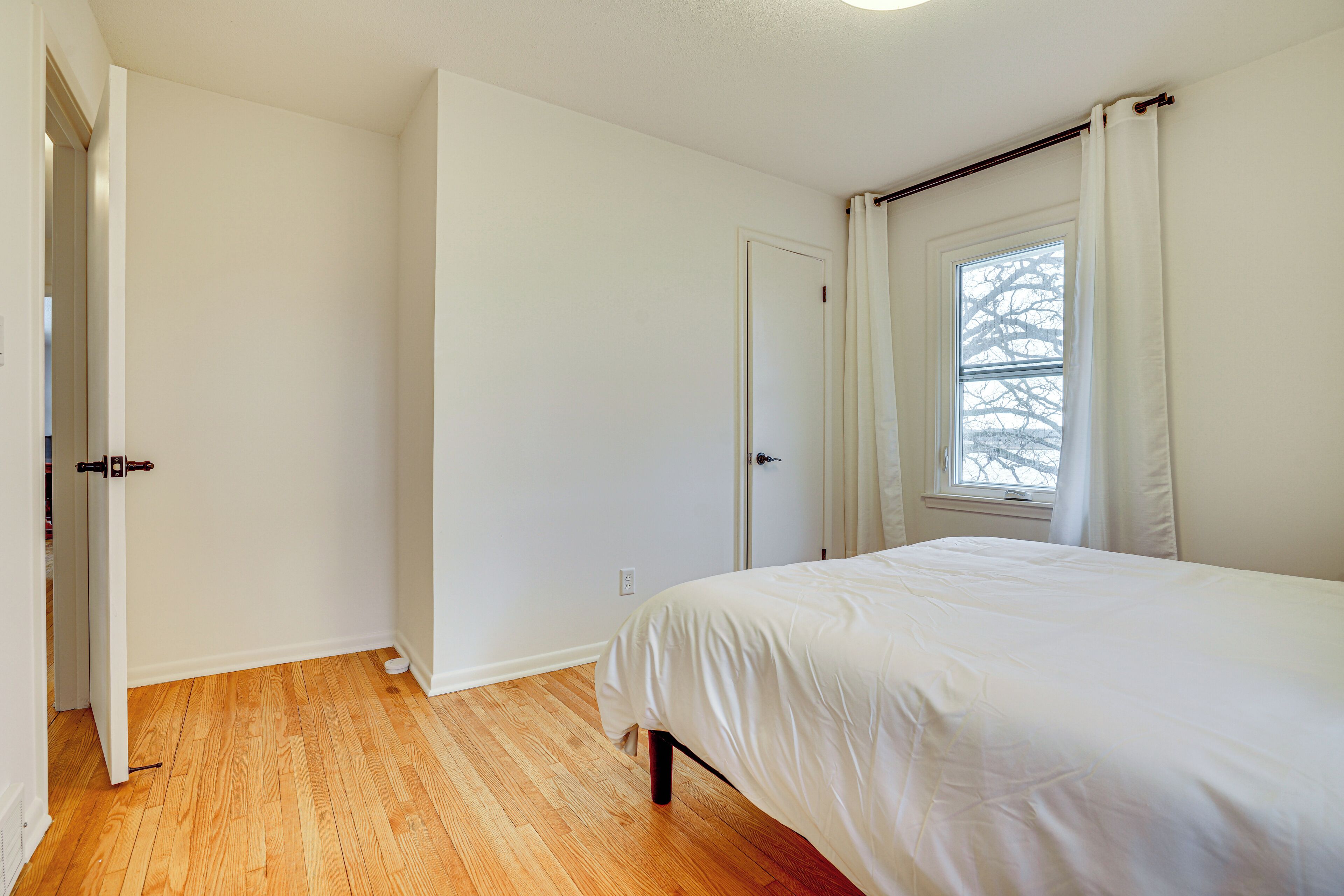 Un dormitorio con piso de madera, una cama con ropa blanca, una ventana con cortinas y una puerta.