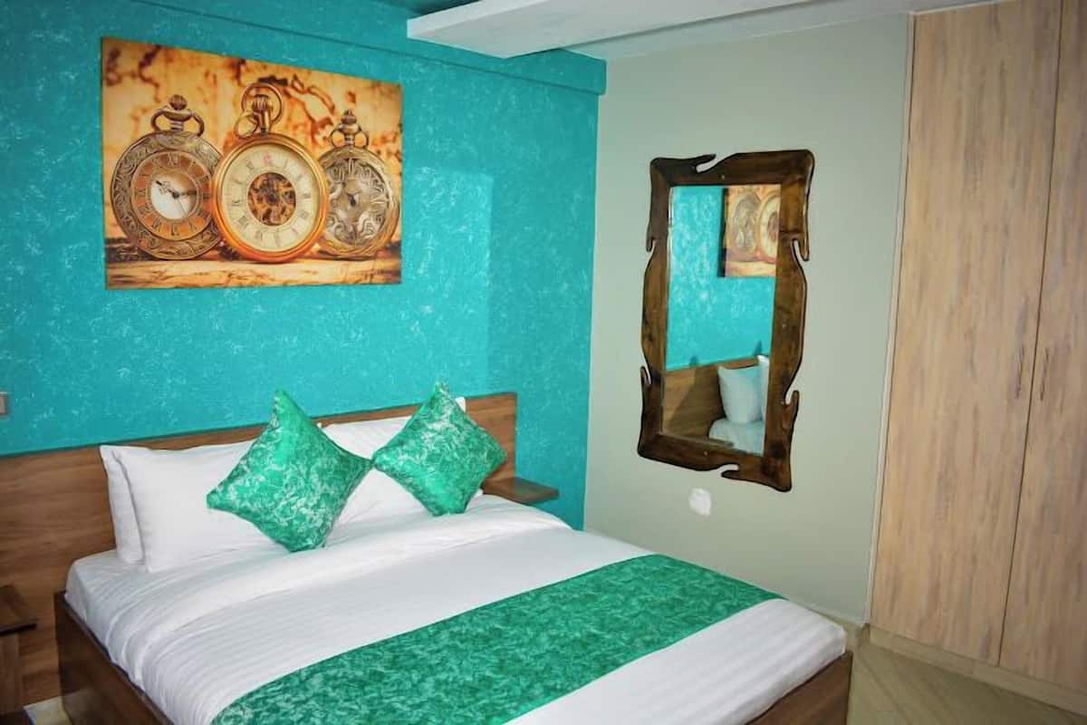 Ein Hotelzimmer mit einem Bett, tealfarbenen Wänden, einem hölzernen Kopfteil, einer grünen Bettdecke und einem Spiegel mit Holzrahmen.
