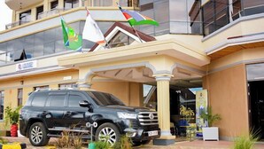 L’entrée d’un hôtel, avec un SUV noir garé devant, des drapeaux et une enseigne indiquant « SALON ».