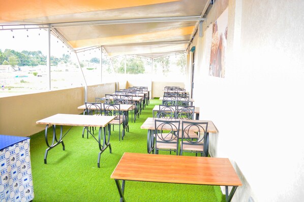 A covered outdoor seating area with tables and chairs, green artificial turf flooring, and a view of the surrounding landscape.