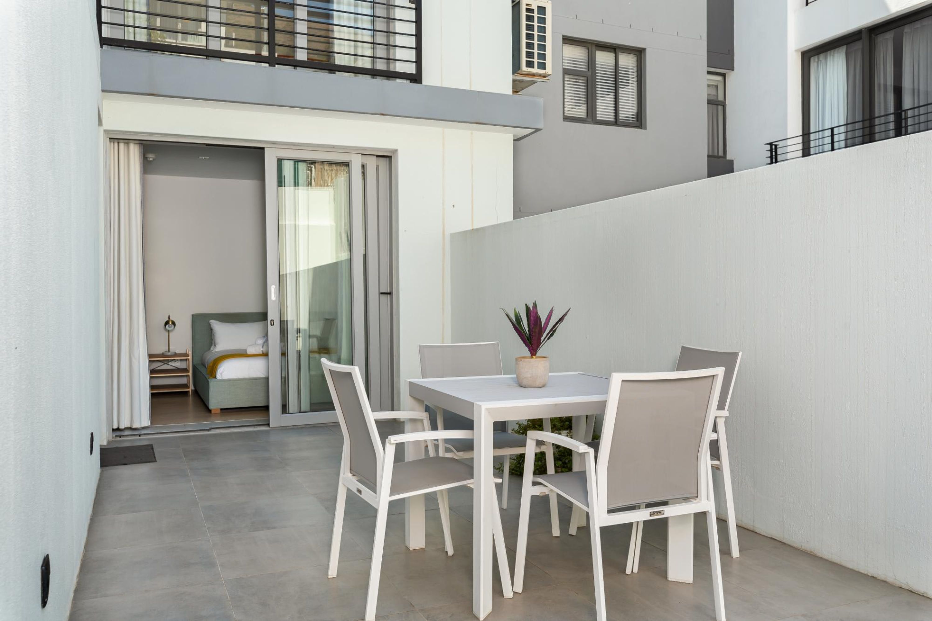 A modern outdoor patio with a white table and chairs, a potted plant, and a sliding glass door leading to a bedroom.