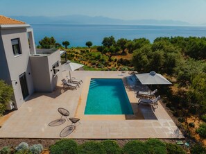 A modern villa with a swimming pool, outdoor seating, and a scenic view of the sea and mountains.