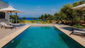 Zona de piscina moderna al aire libre con agua azul cristalina, rodeada de sillones reclinables y sombrillas, con vistas panorámicas al mar y a los árboles.