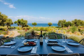 A table set for two with a bottle of wine, plates, cutlery, and a bowl of fruit. The table is outdoors with a view of the sea and trees in the background.