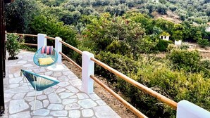 A stone-paved terrace with two blue chairs and a white railing overlooking a hilly landscape with dense vegetation and scattered buildings. - Pergamonto Cottage - Seaview Nest w Private Pool (Skopelos)