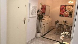 A hallway with a white door, a potted plant, a dining area with a glass table and chairs, and a painting on the wall. - Apartmento en Dama de Noche 1 (Marbella)