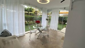 A modern dining area with a glass table and chairs, white curtains, and a view of a garden. - Apartmento en Dama de Noche 1 (Marbella)