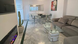 A modern living room with a sofa, a glass coffee table, a fireplace, and a dining area in the background. - Apartmento en Dama de Noche 1 (Marbella)