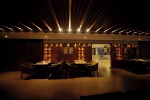 A dimly lit restaurant with a modern interior, featuring a long dining table and chairs, and warm lighting.