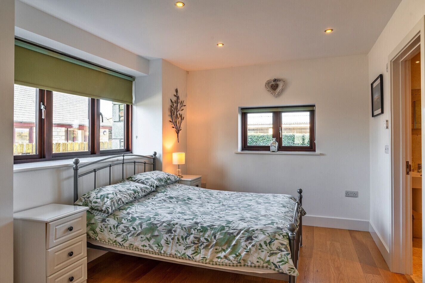A bedroom with a bed, bedside tables, a window with a view, a wall-mounted light, and a heart-shaped decoration.